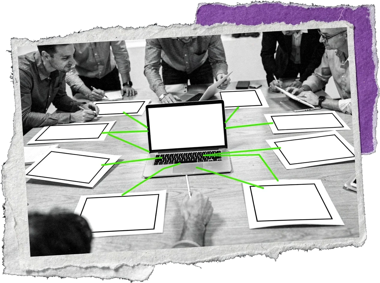 Black-and-white photo of people leaning over a table with a laptop in the centre and blank pages around it, joined to the laptop by green lines.
