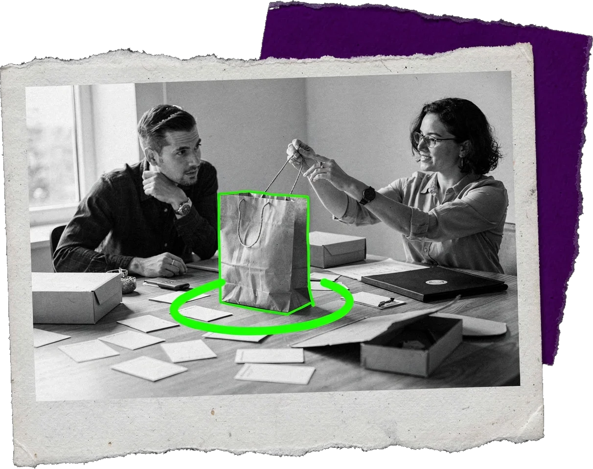 Black-and-white photo of a man and a woman at a table with cardboard boxes and loose sheets, holding up a paper shopping bag; the bag is outlined in green.
