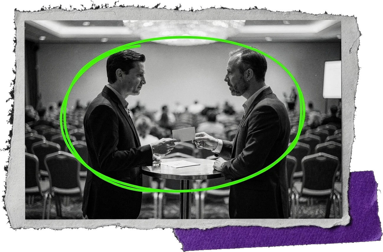Black-and-white photo of two men in suits exchanging a card at a small table in a conference hall, circled in green.