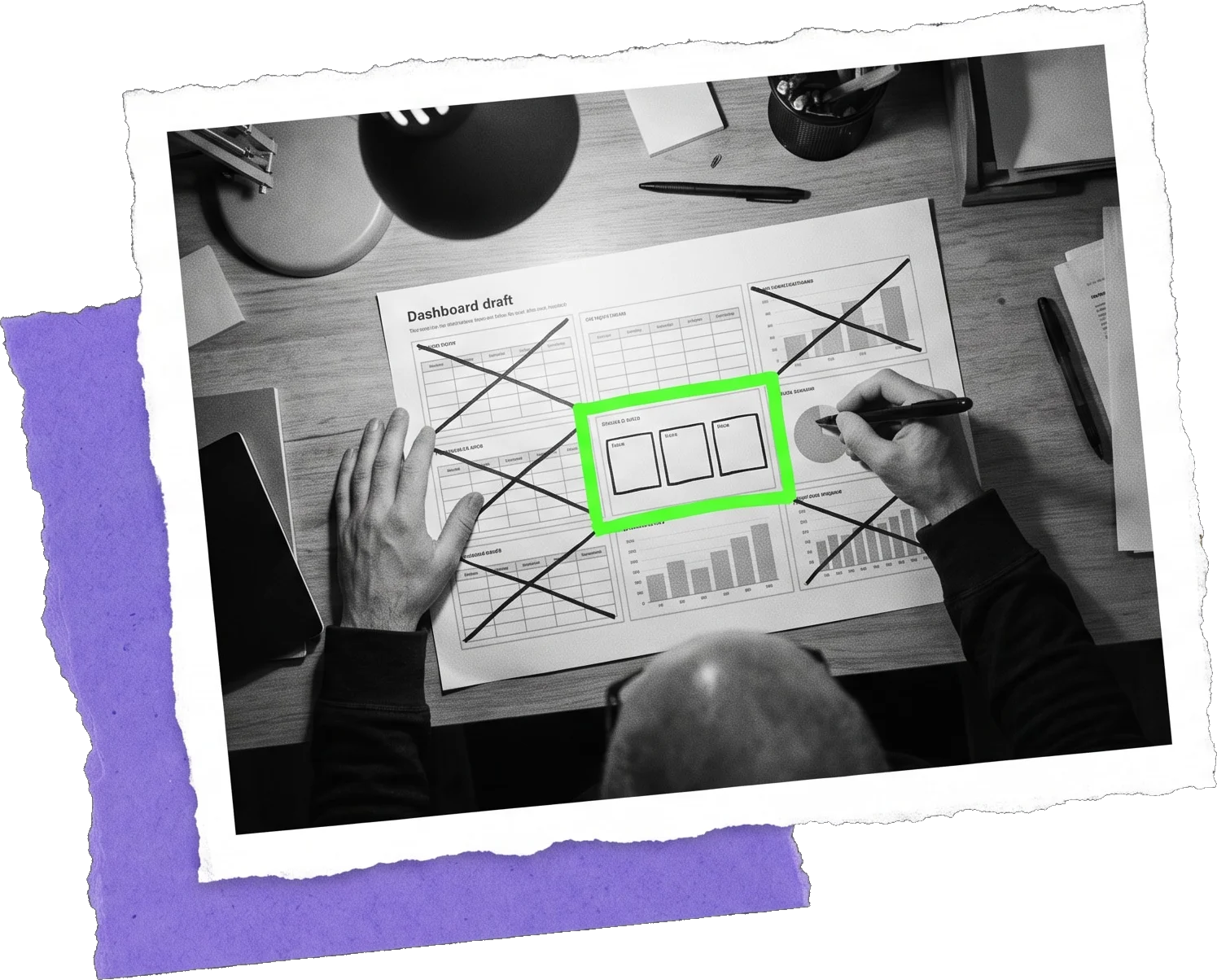 Black-and-white photo from above of a paper dashboard draft with most tiles crossed out and three tiles highlighted in green; a hand holds a pen over it.